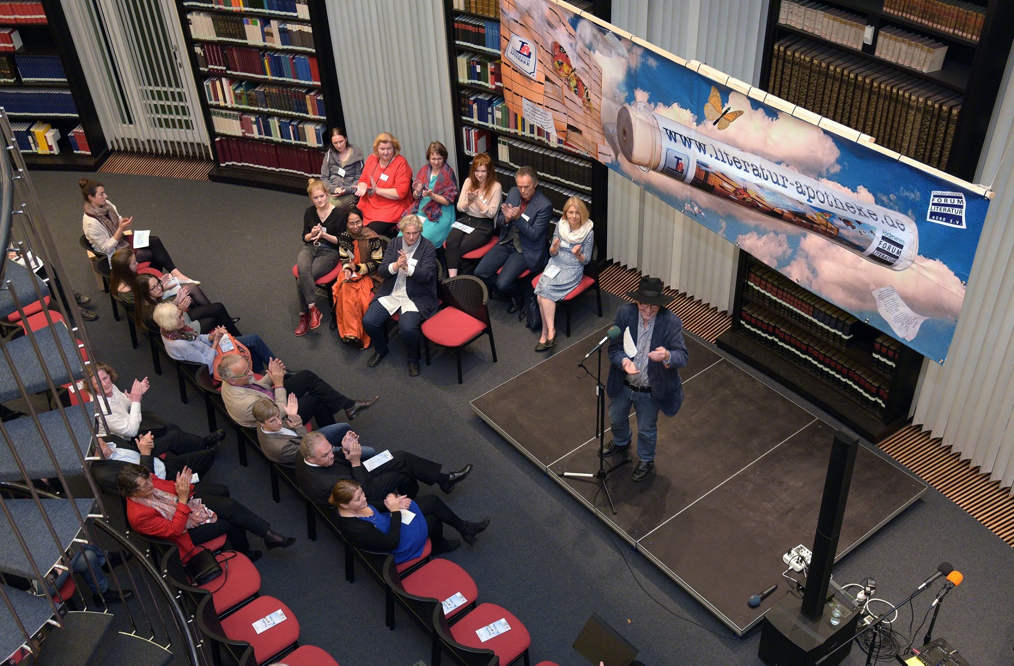 Jo Köhler steht bei einer Veranstaltung im Rahmen der Literatur-Apotheke in einer Bibliothek auf der Bühne, das Publikum applaudiert
