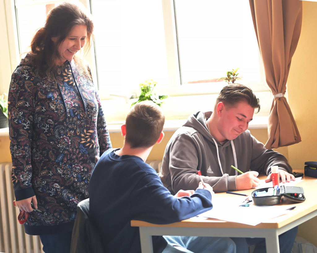 Die Lehrerin schaut zwei jungen Männern über die Schulter und lacht freundlich.