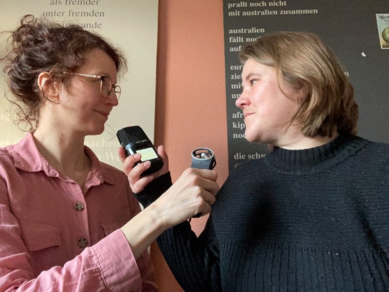 Mitarbeiterinnen Lucie Kling und Rachel Bleiber halten sich gegenseitig ein Mikrofon vor den Mund.