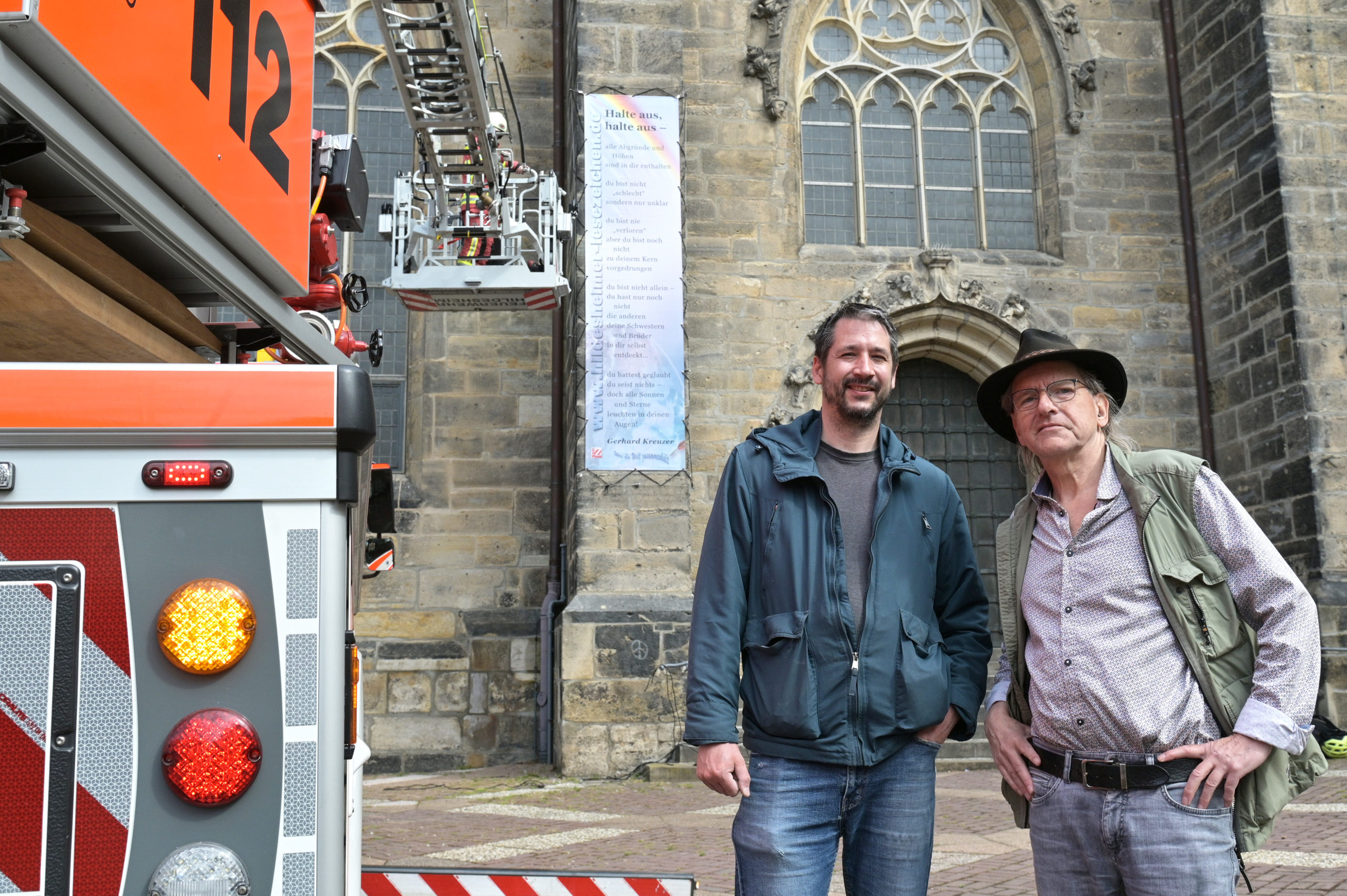 Der Monteur Benjamin Spitzbart und Jo Köhler stehen vor der Andreaskirche. An der Kirchenwand ist ein großes Gedichtbanner angebracht. Im Hintergrund ein Feuerwehrmann auf einem Leiterwagen.