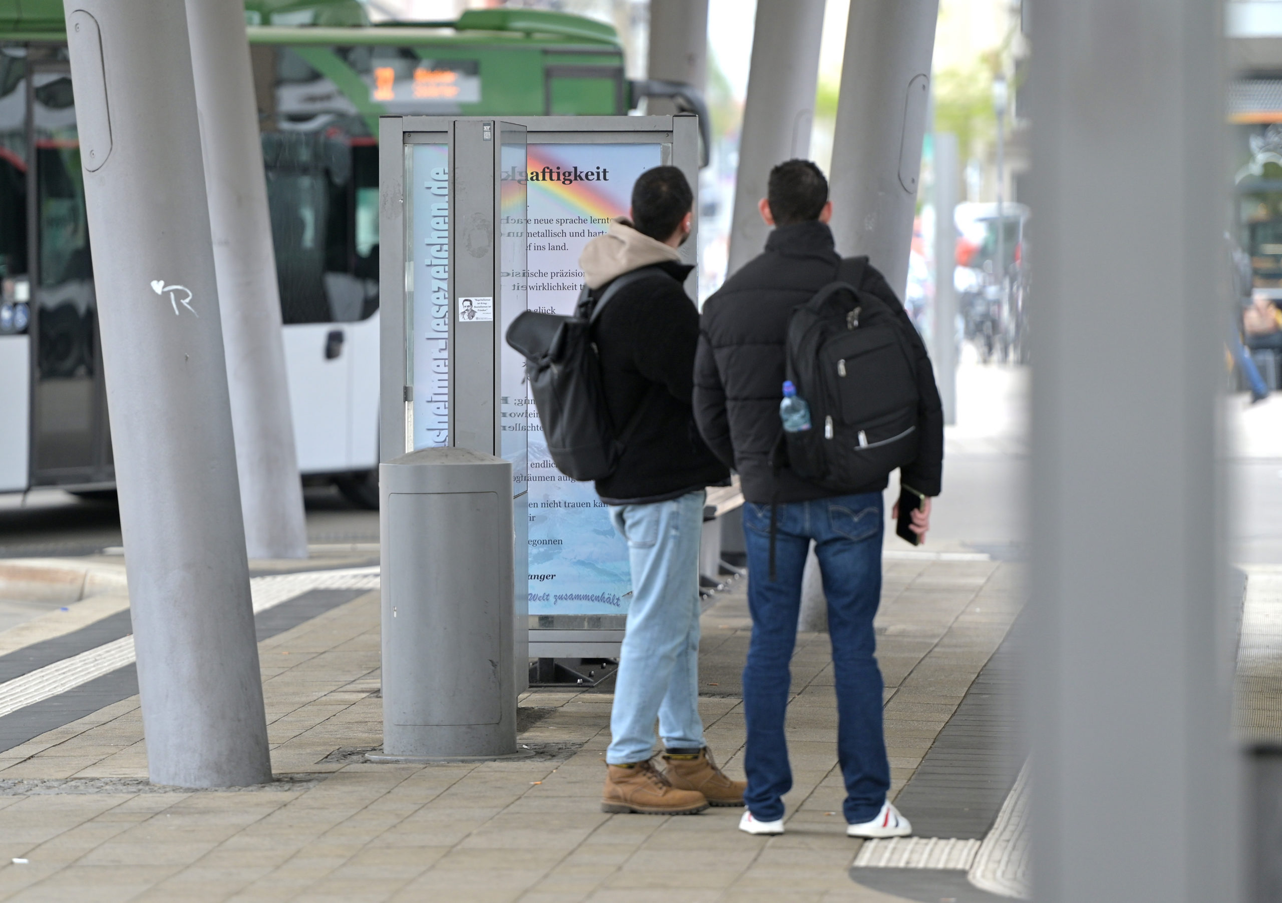 Zwei junge Männer stehen am Busbahnhof vor einem Gedichtbanner.