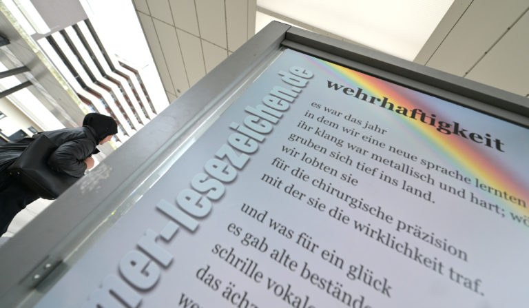 Das Gedicht "wehrhaftigkeit" am Busbahnhof. In der linken Ecke eine junge Frau, die ein Kopftuch trägt.