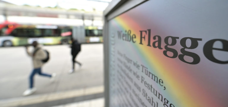 Das Gedicht "Weiße Flagge" am Busbahnhof. Im Hintergrund rennen zwei Männer über die Fahrbahn.