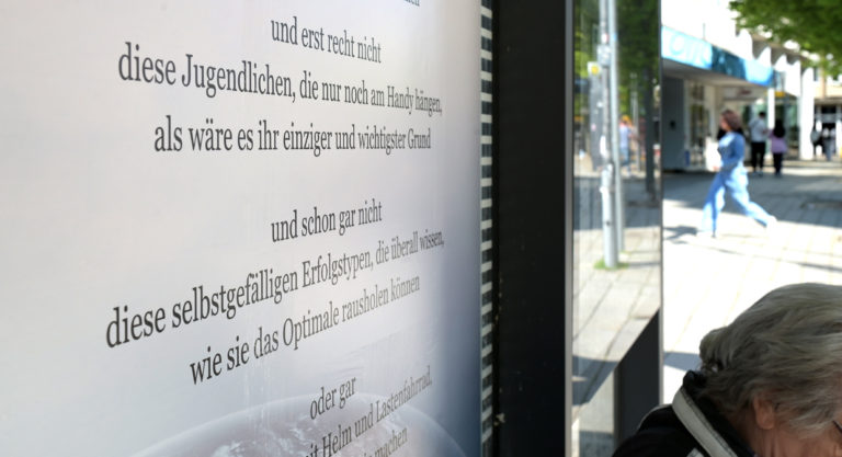 Ein Gedicht hängt an einer Bushaltestelle. Davor sitzt eine ältere Dame.