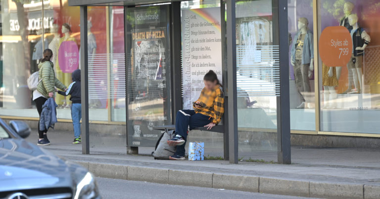 Eine Frau sitzt an der Bushaltestelle und schaut auf ihr Handy. Hinter ihr hängt ein Gebet.